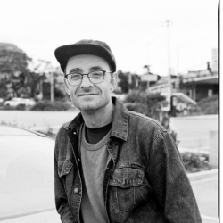 Lecturer Sean Sanford, wearing a hat, glasses and a denim jacket, with a blurry background